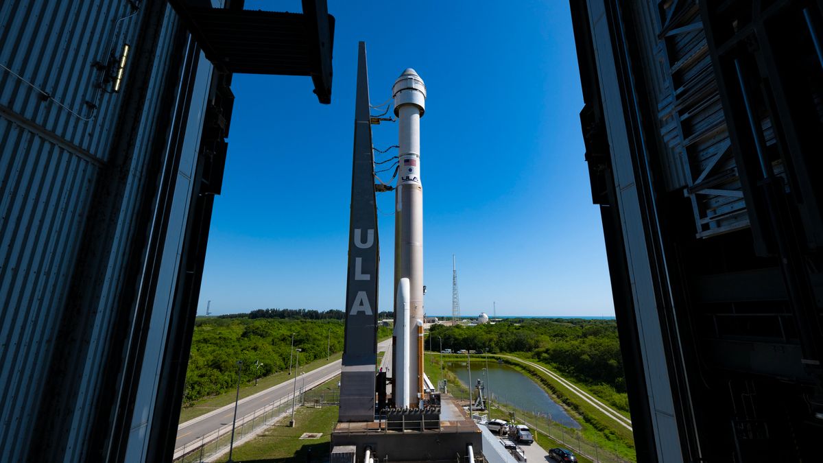 a brown and white rocket carrying a white space capsule rolls out of an assembly building toward the launch pad