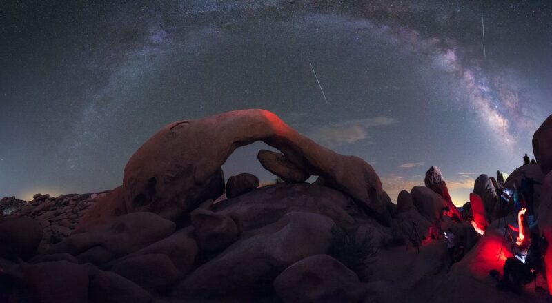 A rocky arc sits above a field of jagged rocks. In the dark blue sky behind, an arc of stars and dark cloudy dust stretches across the frame, with two white streaks shooting across the sky.