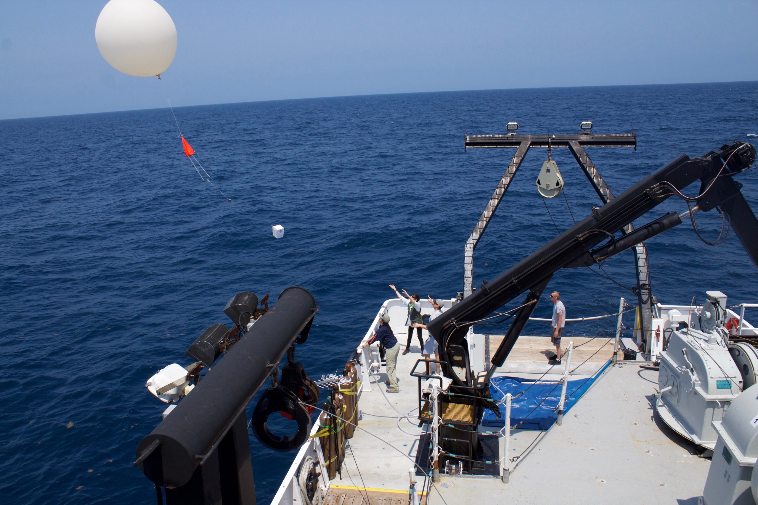 In the foreground of the image in the bottom right corner of the image is a portion of a boat. The deck of the boat is a gray color and has several large pieces of equipment on it including pully systems. There are four people standing on the deck of the boat. In the background is the deep blue ocean, which looks flat, and meets up with the light blue sky at the horizon. In the top left corner of the image is a large white balloon, which has just been released from the boat.