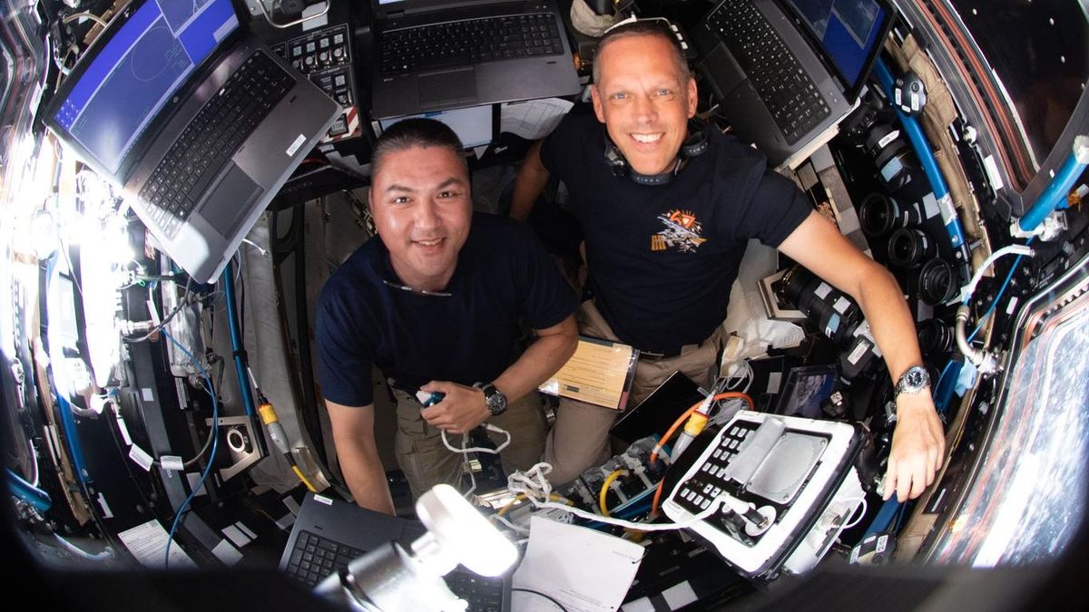 an overhead shot of two astronauts smiling in a small windowed room of the international space station, surrounded by computers and a window